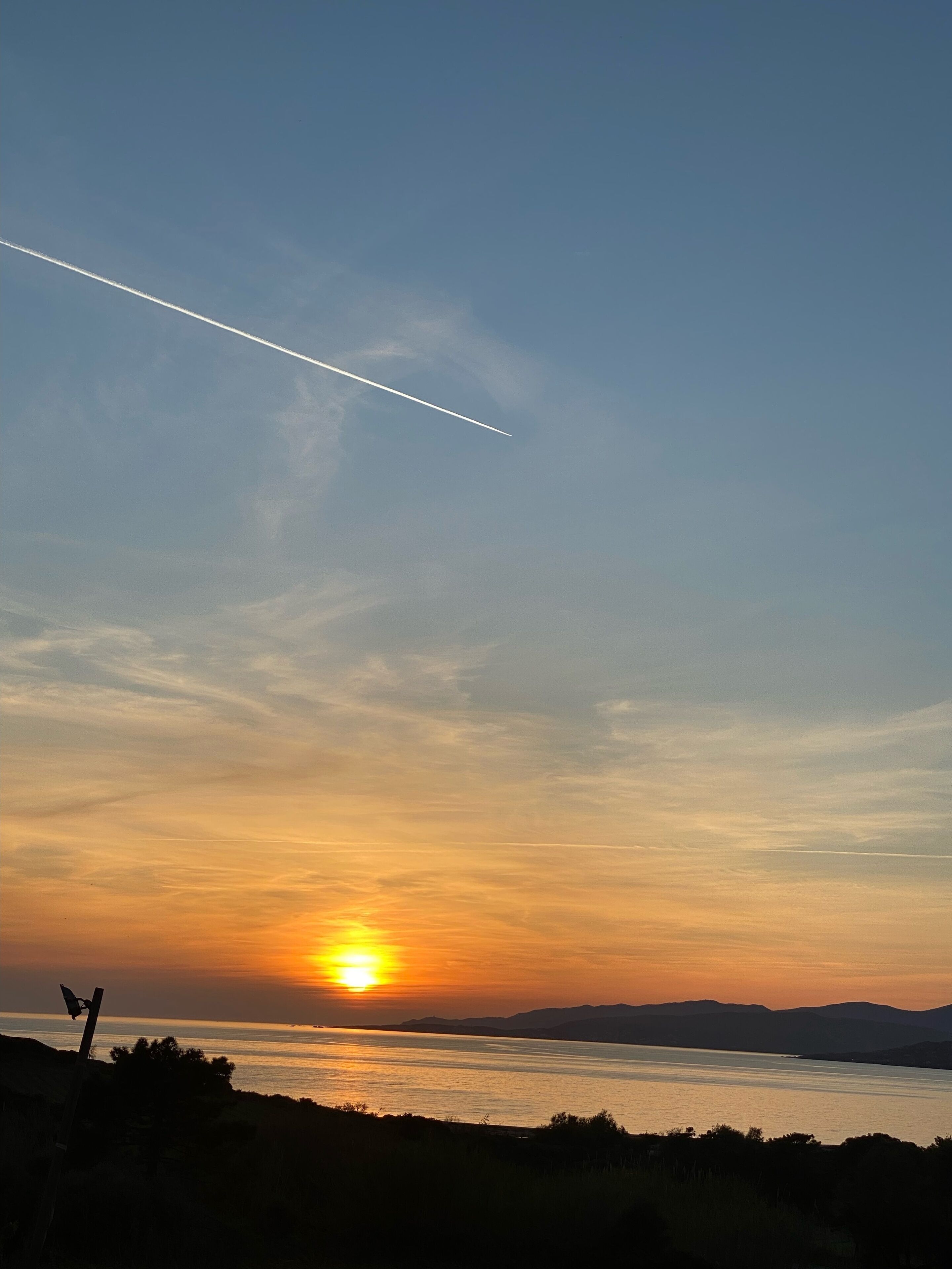 A sunset over a body of water with a clear sky and a contrail.