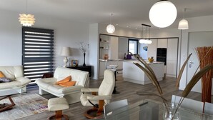 Una sala moderna con un sofá, sillones y una mesa de centro de vidrio. Se ve un área de cocina con una mesa de comedor y sillas al fondo.