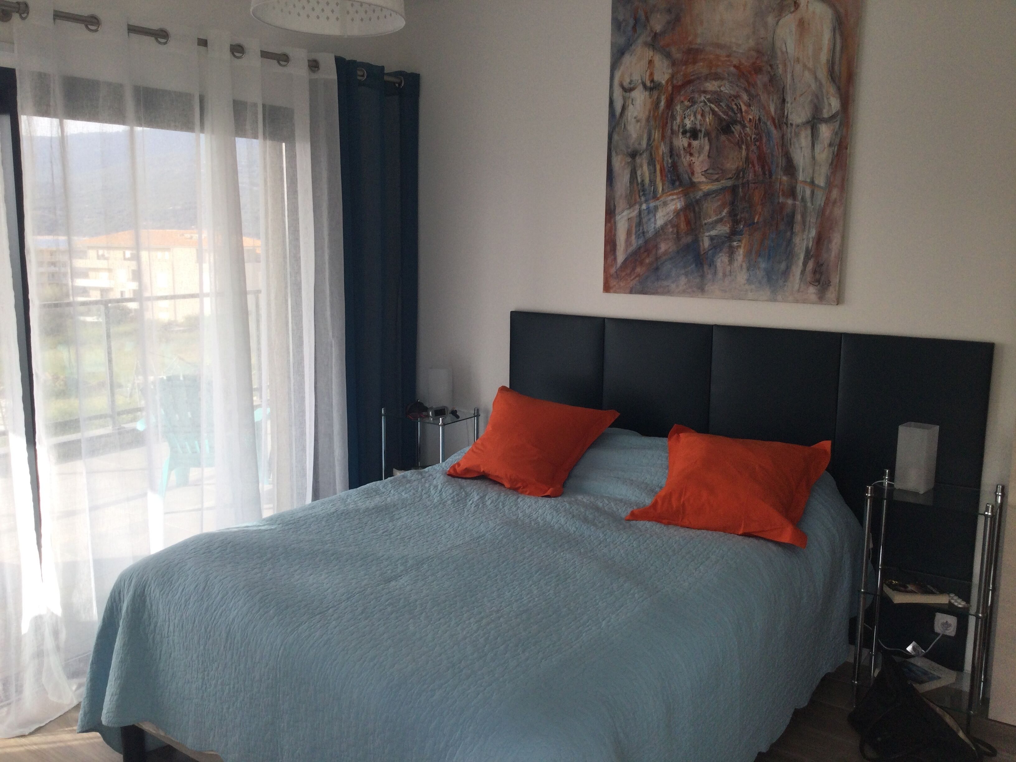 Une chambre à coucher avec un lit, un tableau au mur, une fenêtre avec des rideaux et une table de chevet.