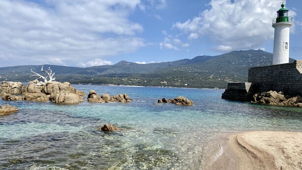 Escena costera con un faro, agua cristalina, afloramientos rocosos y una montaña al fondo.