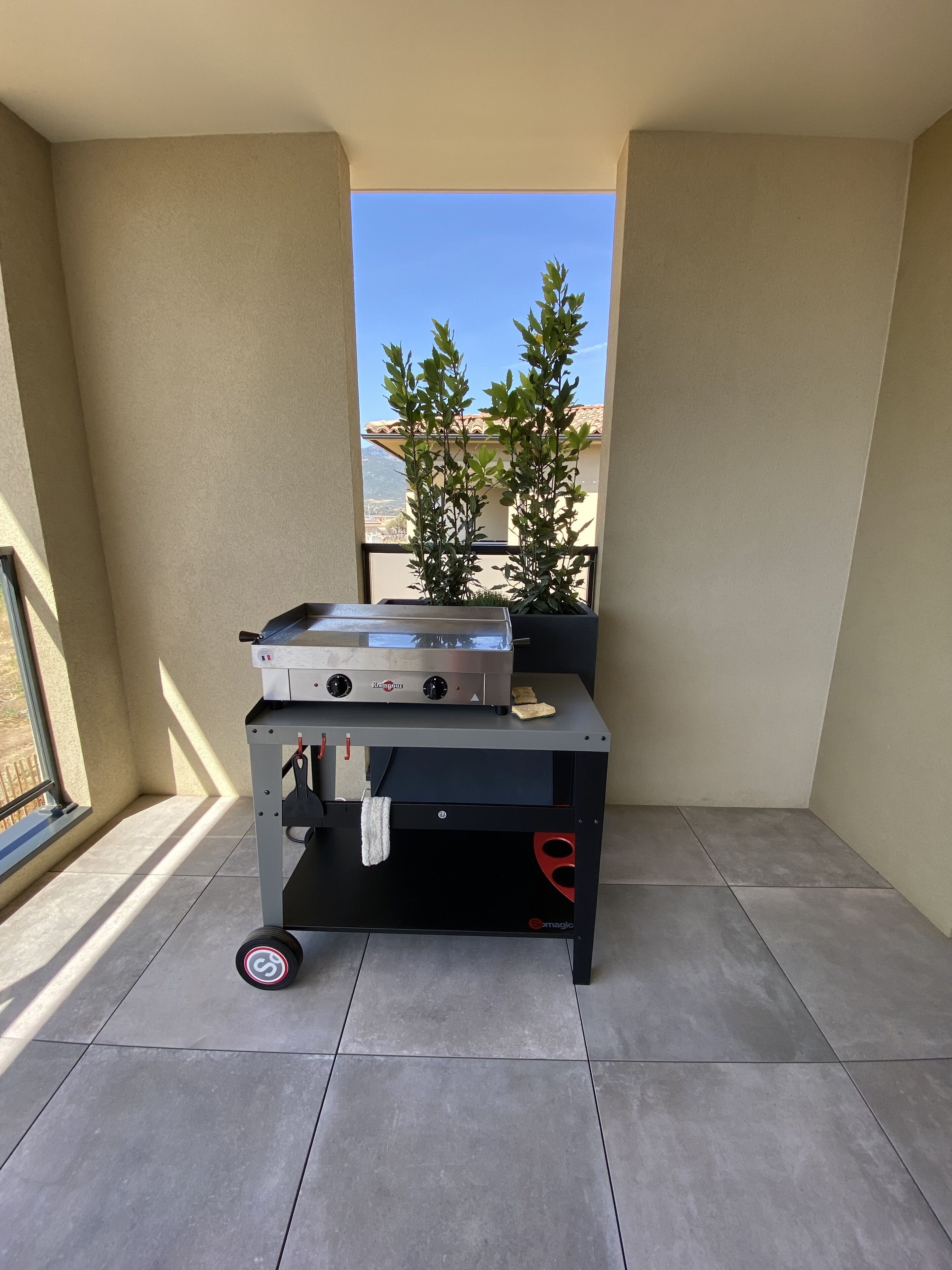 A compact outdoor cooking area with a portable grill and a small table, situated on a balcony with a view of a building and trees.
