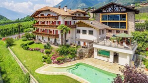 A two-story house with a swimming pool, surrounded by lush greenery and mountains.