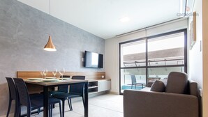 A modern living space with a dining area, a sofa, a TV mounted on the wall, and a large window offering a view. - Porto Blue ao mar de Porto G - Carpediem (Ipojuca)
