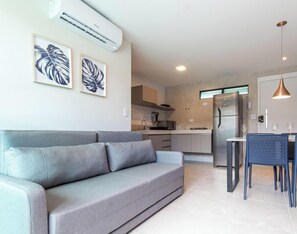 A modern living room with a grey sofa, a kitchen with a stainless steel refrigerator, and a dining area with a table and chairs. - Porto Blue ao mar de Porto G - Carpediem (Ipojuca)