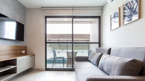 A modern living room with a sofa, a flat-screen TV, and a balcony with a view. - Porto Blue ao mar de Porto G - Carpediem (Ipojuca)