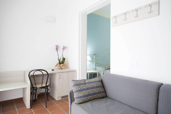 Una habitación pequeña con un escritorio blanco, una silla negra, un cojín estampado sobre un sofá gris, una planta pequeña y un panel de ganchos en la pared.