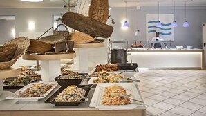 A buffet with various dishes, a counter with a person behind it, and hanging pendant lights. - Blu Hotel Laconia (Arzachena)