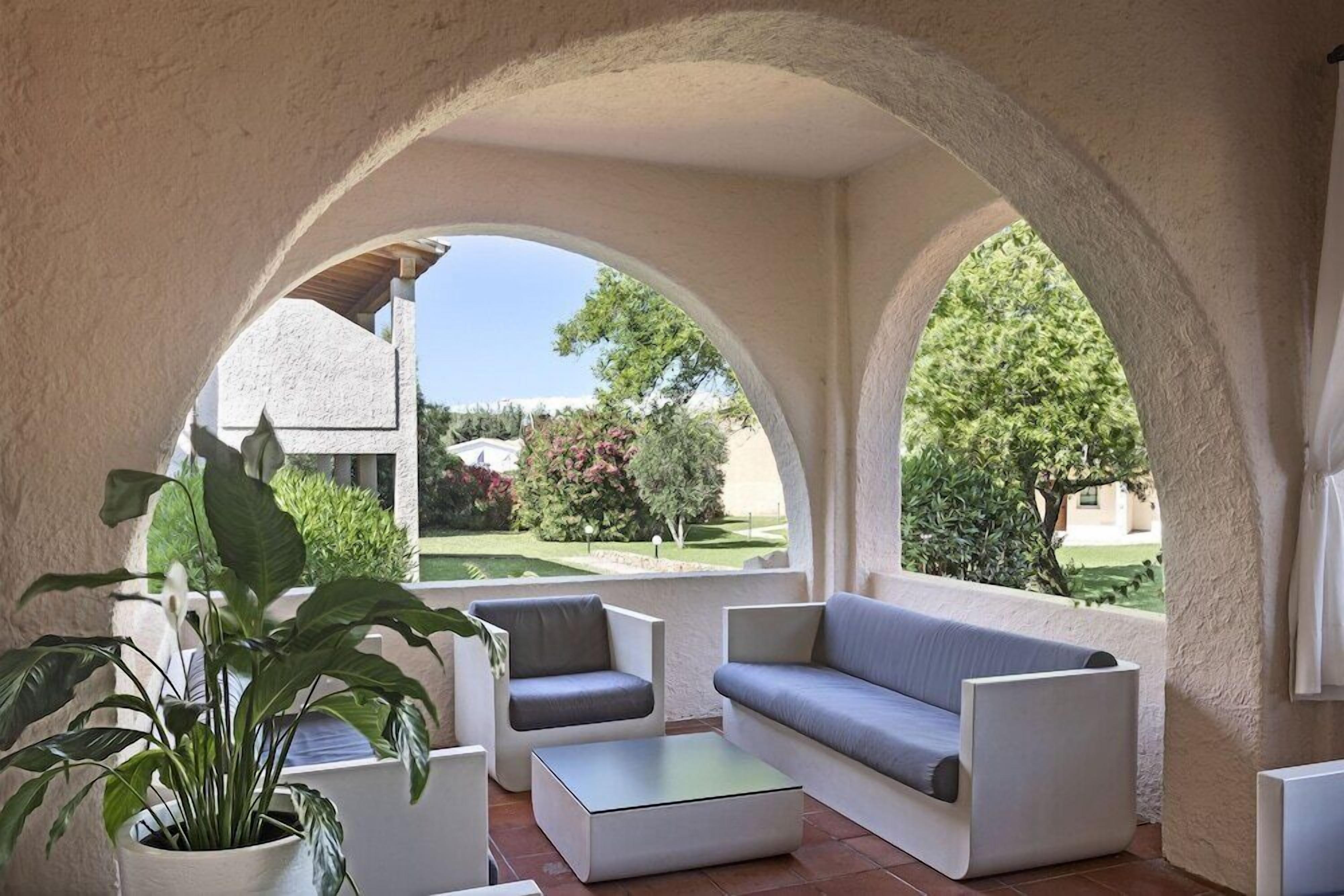 Area salotto esterna con divano, sedie e tavolino, incorniciata da archi e piante in vaso.
