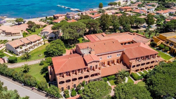 An aerial view of a coastal town with a prominent building featuring a red-tiled roof and multiple arches, surrounded by lush greenery and a clear blue sea. - Blu Hotel Laconia (Arzachena)