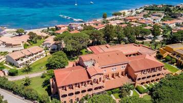 Una vista aerea di una cittadina costiera con un edificio importante caratterizzato da un tetto in tegole rosse e più archi, circondato da vegetazione lussureggiante e un mare cristallino.