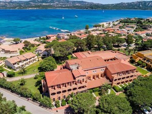 Vista aérea de un pueblo costero con un edificio destacado, techo de tejas rojas y múltiples arcos, rodeado de abundante vegetación y un mar azul cristalino.