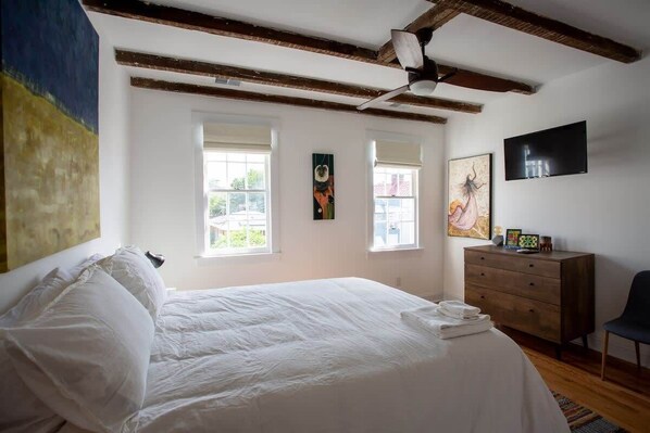 A bedroom with a large bed, two windows, a ceiling fan, a TV, and a dresser.