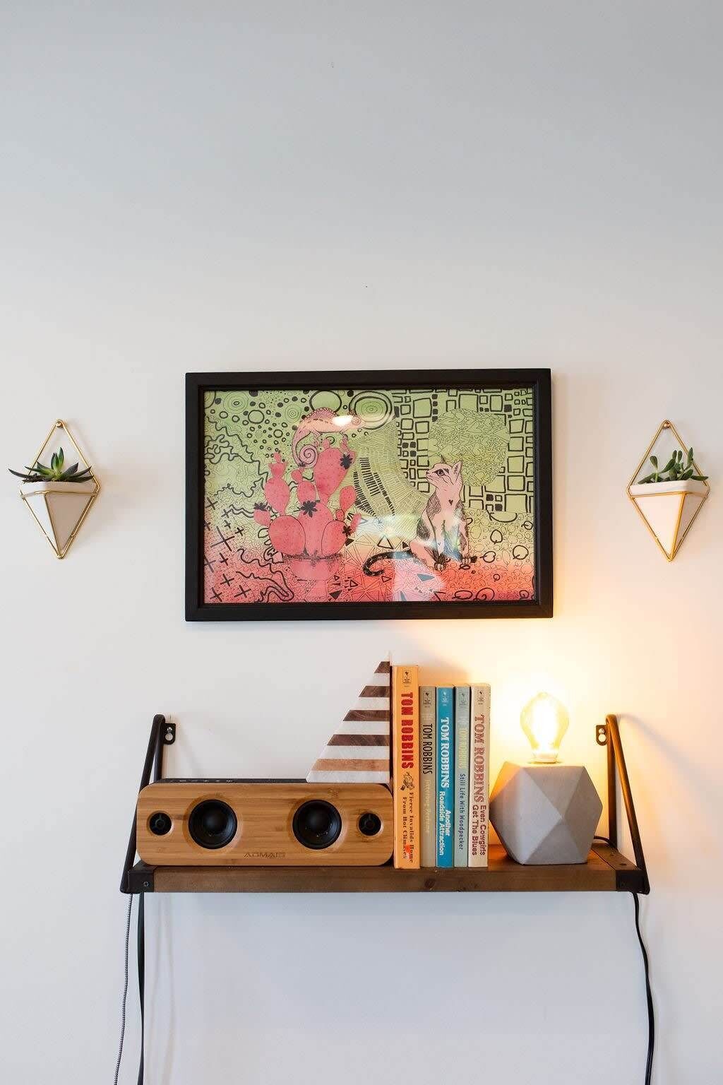 Una obra de arte enmarco con diseños abstractos y dos pequeñas macetas a cada lado. Debajo, un estante de madera sostiene libros y una lámpara.