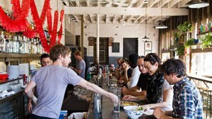 Un espace bar où des clients sont assis à des tables, un barman sert des boissons et des étagères sont remplies de bouteilles et de verres.