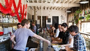 Un’area bar con clienti seduti ai tavoli, un barista al bancone e scaffali pieni di bottiglie e bicchieri.