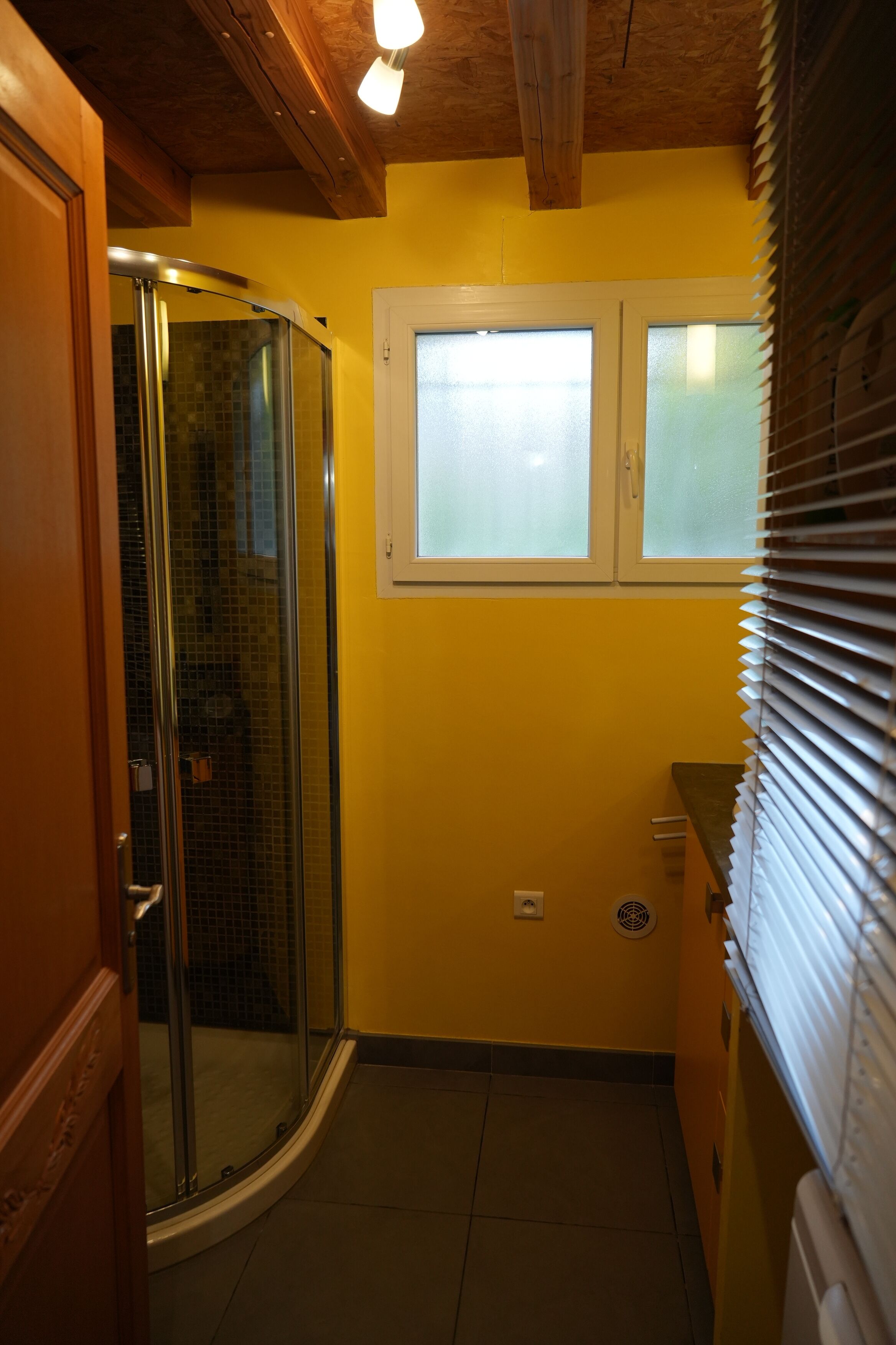 A bathroom with a glass shower enclosure, a window with blinds, and a yellow wall.