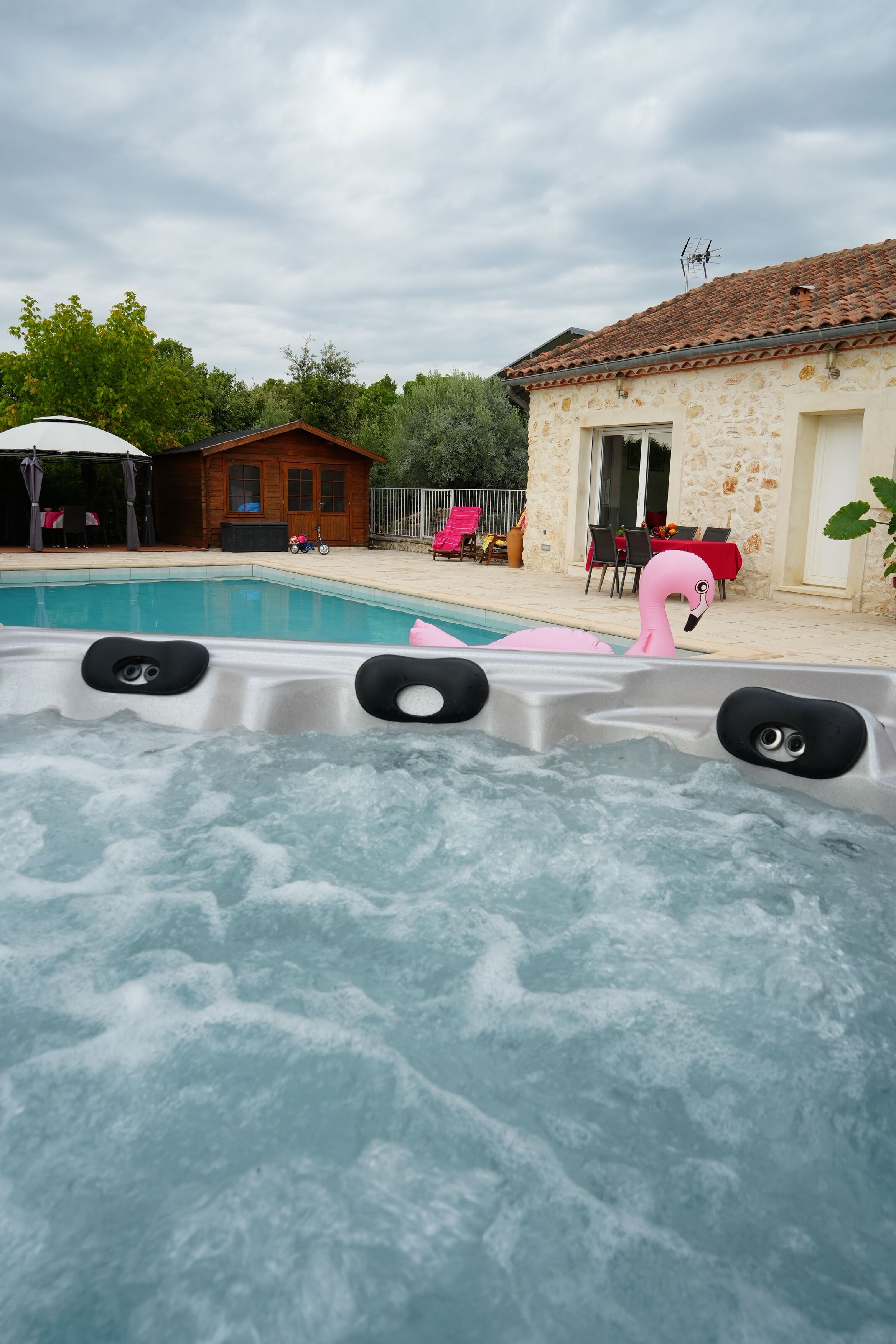 Un bain à remous avec vue sur une piscine, un chalet en bois et un bâtiment en pierre avec un toit en tuiles.