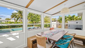 A spacious dining area with a large table, wicker chairs, and a view of a swimming pool and palm trees. - Rove Travel | Sunny Vista | Heated Pool & Firepit (West Palm Beach)