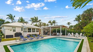 A swimming pool with lounge chairs, a covered patio, and palm trees. - Rove Travel | Sunny Vista | Heated Pool & Firepit (West Palm Beach)