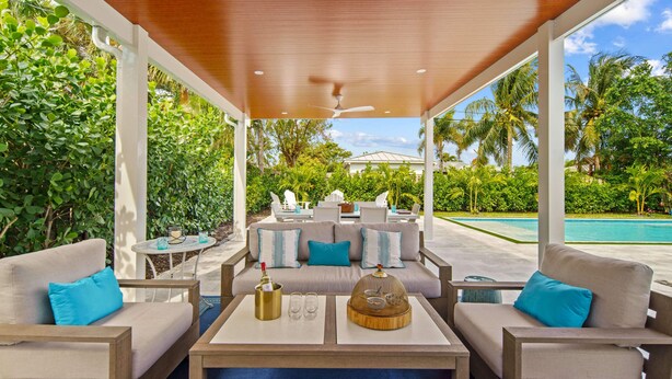 A covered outdoor seating area with sofas and tables, overlooking a pool and lush greenery. - Rove Travel | Sunny Vista | Heated Pool & Firepit (West Palm Beach)