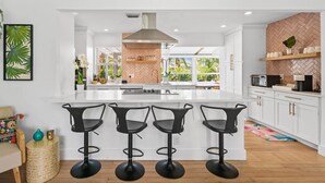 A modern kitchen with a central island, three black bar stools, white cabinetry, and a herringbone-patterned backsplash. - Rove Travel | Sunny Vista | Heated Pool & Firepit (West Palm Beach)