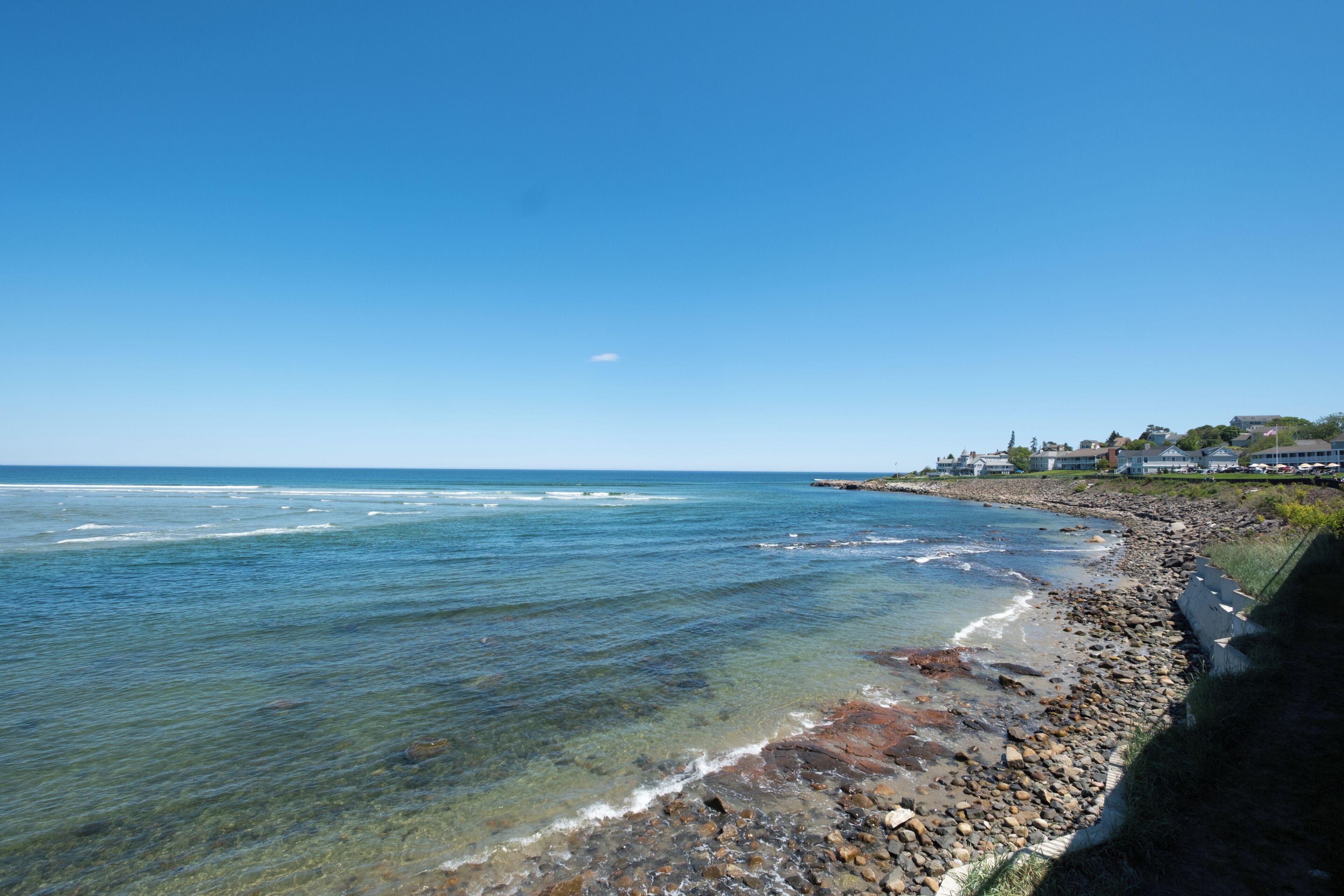 A coastal view with clear water, a rocky shoreline, and a row of houses in the background.