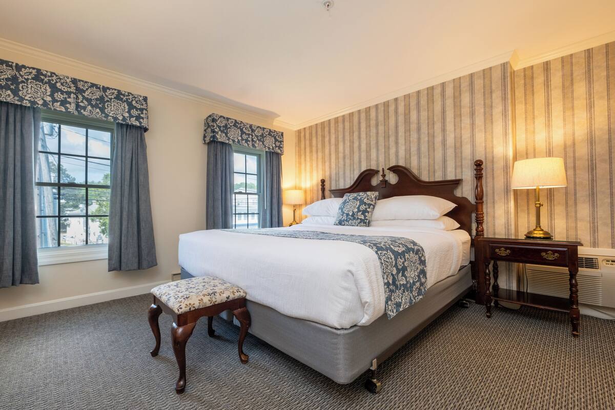 A hotel room with a large bed, two bedside lamps, a wooden nightstand, a bench, and a window with curtains.
