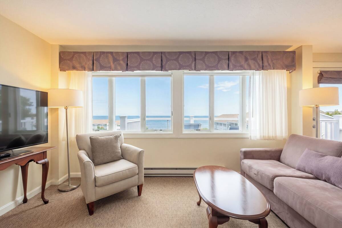 A hotel room with a sofa, armchair, TV, and a view of the sea.