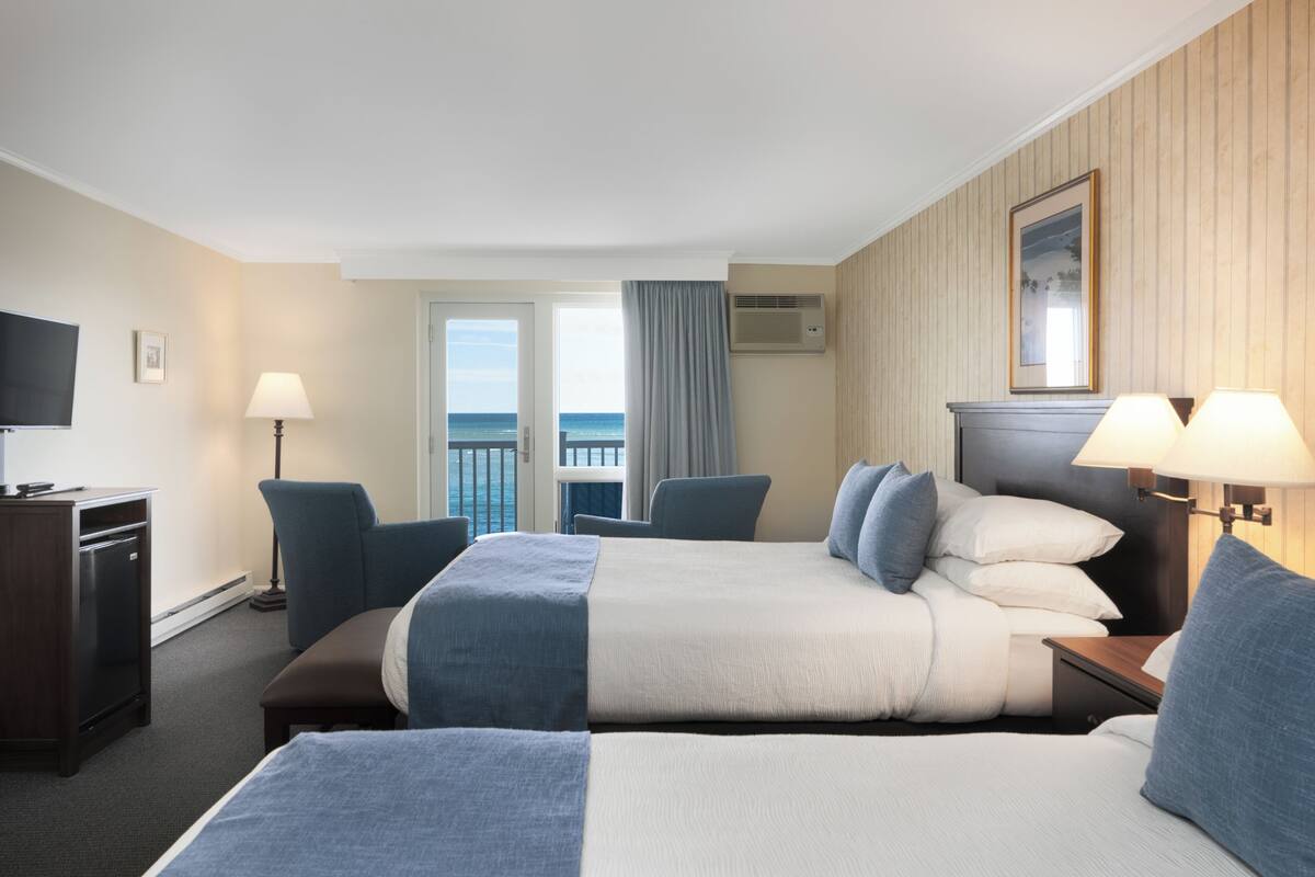A hotel room with two beds, a desk, a TV, a window with a view, and a wall-mounted air conditioner.