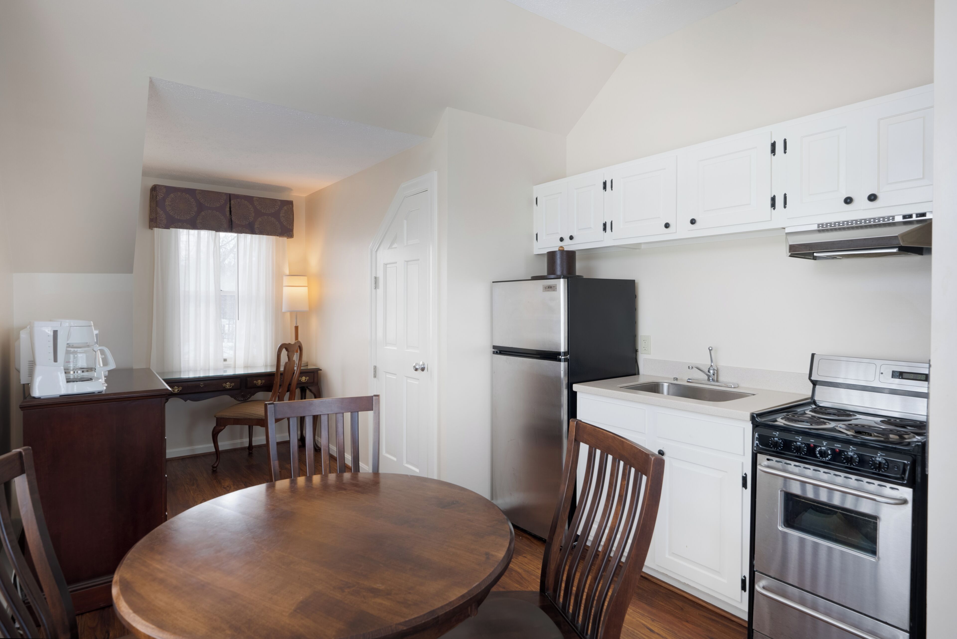 A compact kitchen with white cabinets, a stainless steel refrigerator, a stove, and a dining table with chairs.