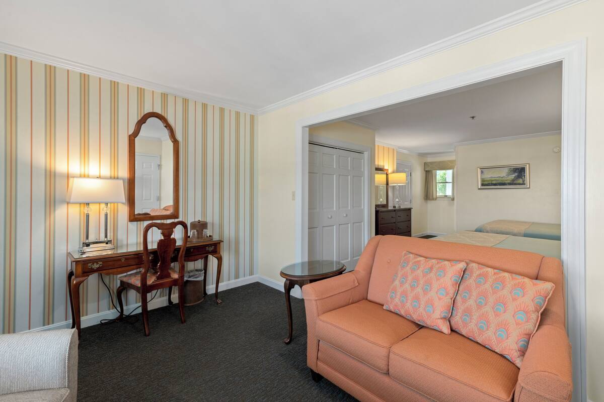 A hotel room with a desk, chair, and sofa. There is a mirror above the desk, a lamp on the desk, and a painting on the wall.