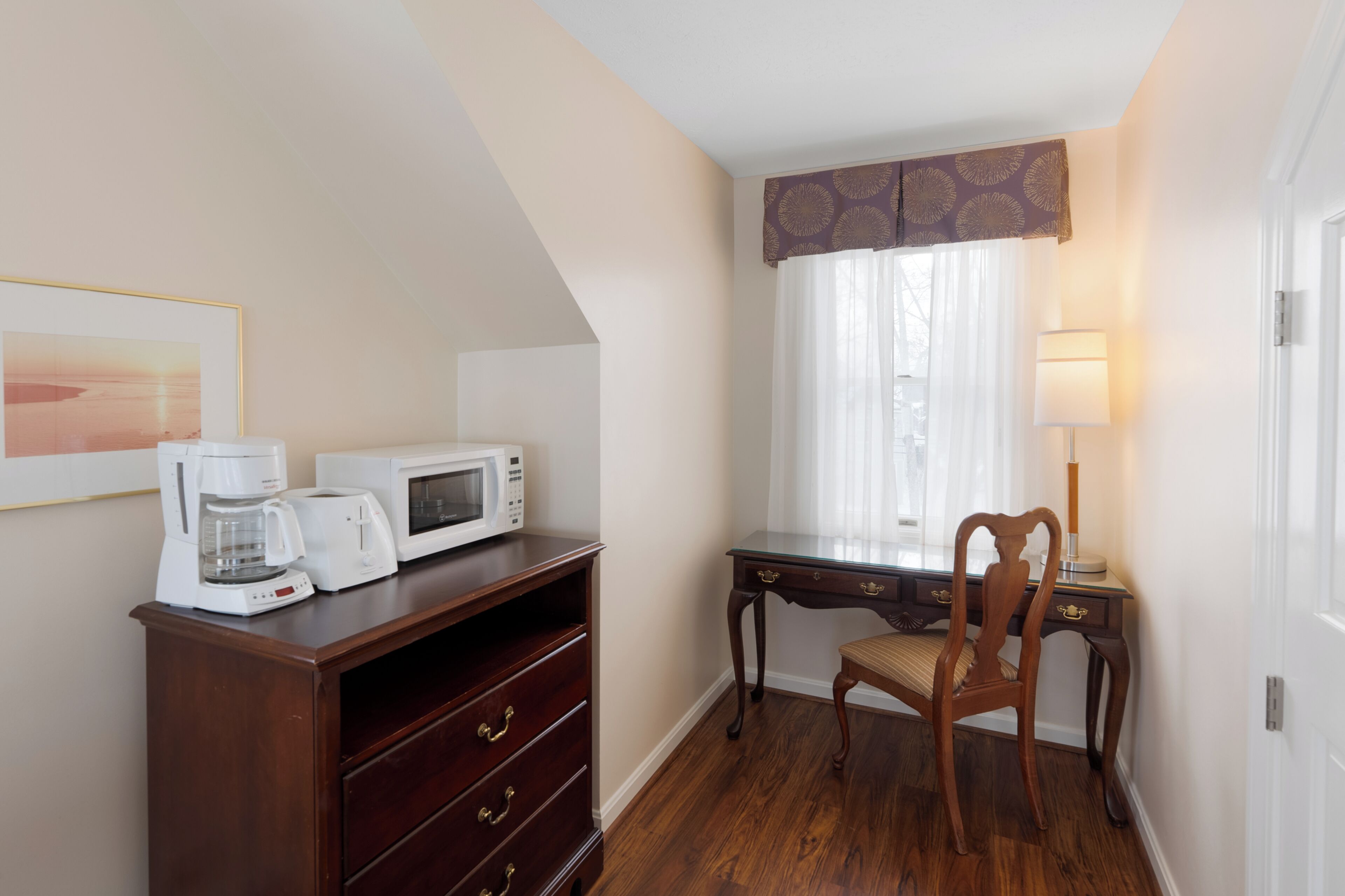 A hotel room with a wooden desk, a chair, a coffee maker, a microwave, and a lamp.