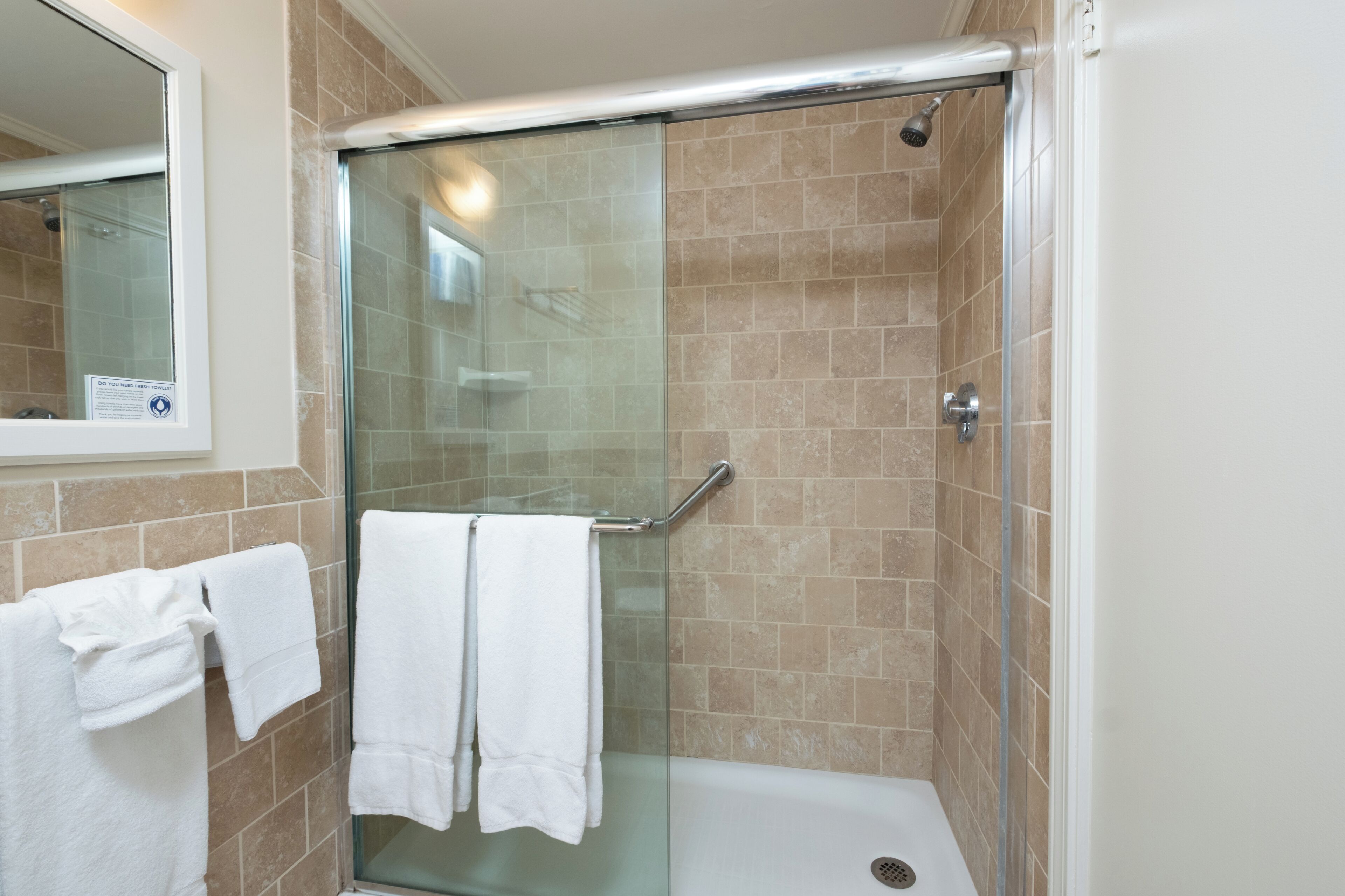 Um chuveiro com paredes de vidro, uma banheira branca, azulejos bege e uma porta de vidro para o chuveiro.