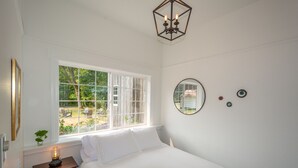A bedroom with a large window, a bed, a bedside table with a lamp, a round mirror, and a geometric ceiling light.