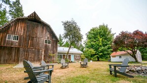 A rustic barn, a small white house, and several Adirondack chairs in a grassy yard.