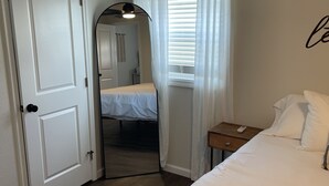 A bedroom with a bed, a nightstand, a mirror, and a door leading to another room. - Centrally Located - Newly Built - Walk To The Beach! (Avon)