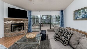 A living room with a brick fireplace, a glass coffee table, a sofa with patterned cushions, and a large window with a view of a garden.