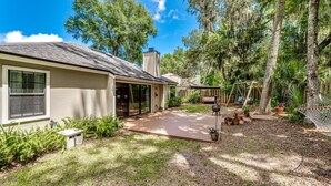 A house with a covered patio, a wooden deck, and a fenced yard with trees and plants.