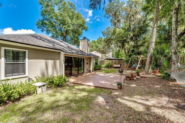 A house with a covered patio, a wooden deck, and a fenced yard with trees and plants.