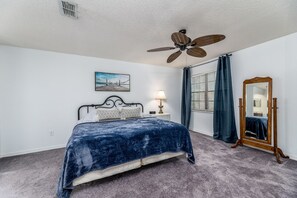 A bedroom with a bed, a ceiling fan, a nightstand with a lamp, a large mirror, and a window with curtains.