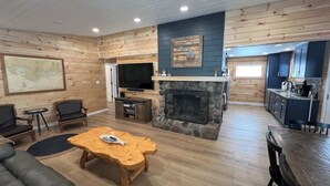 A spacious living room with a stone fireplace, a television, and a kitchen area with dark cabinets. - 'Lakeside' Cottage with porch and sandy beach (Cedarville)