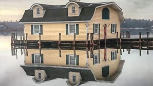 A house with a symmetrical design, featuring a central entrance and two side windows, surrounded by a dock extending into a calm body of water. - 'Lakeside' Cottage with porch and sandy beach (Cedarville)