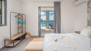 Een moderne slaapkamer met een groot bed, een houten bankje, een metalen plank en uitzicht op de oceaan door een glazen deur.