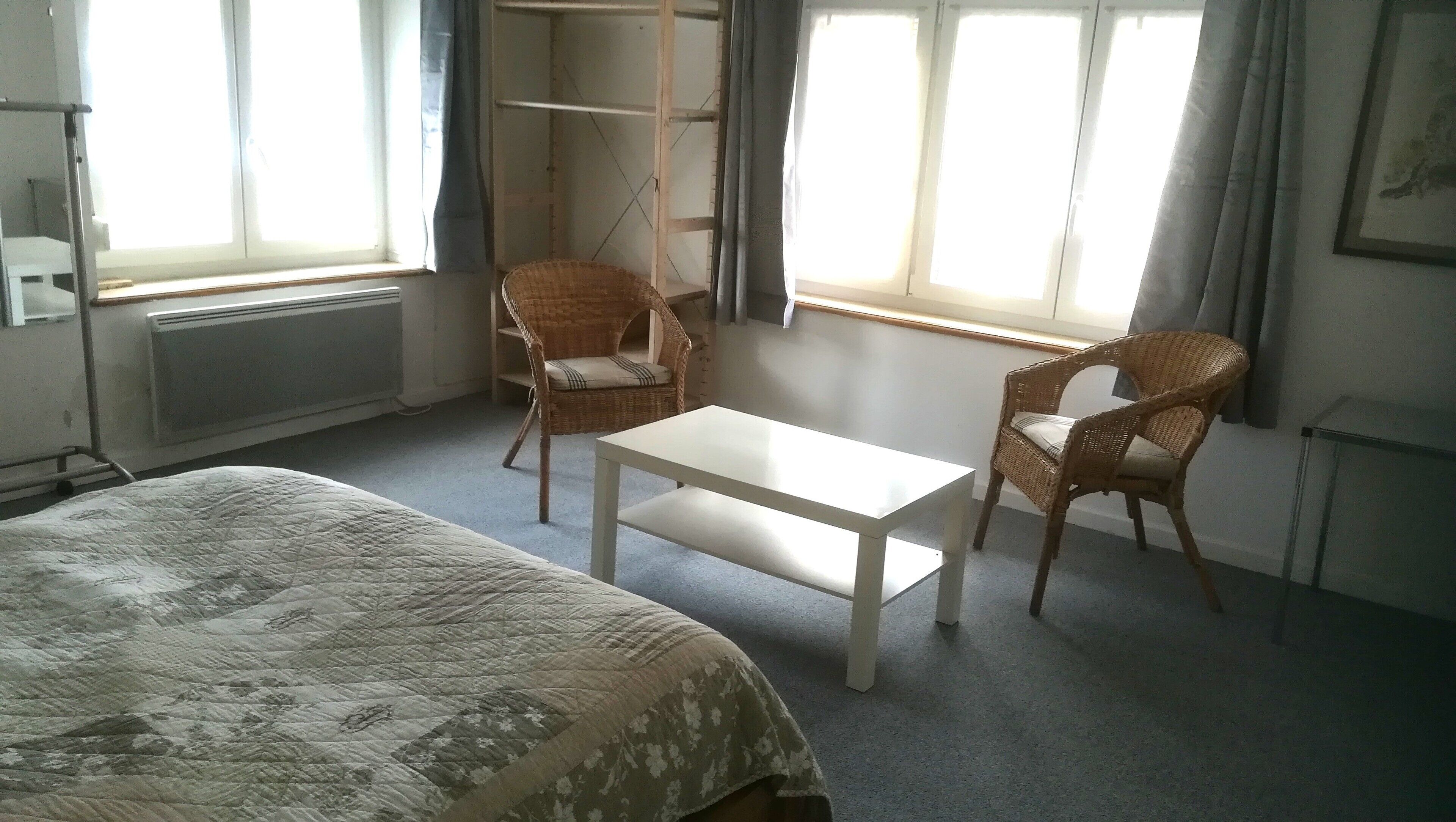 Ein Hotelzimmer mit einem Bett, einem kleinen Tisch, zwei Korbstühlen, einem Kleiderschrank und einem Fenster mit Vorhängen.