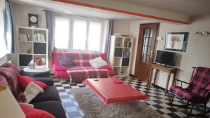 Un salon avec un sol à carreaux, un canapé rouge, un canapé noir, une table basse rouge et un fauteuil rouge. On y trouve également deux étagères, un téléviseur et une porte.