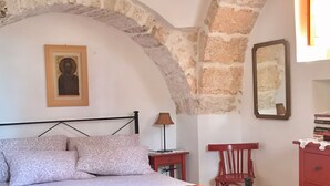 Een slaapkamer met een bed, een stoel, een klein tafeltje, een lamp en een dressoir.