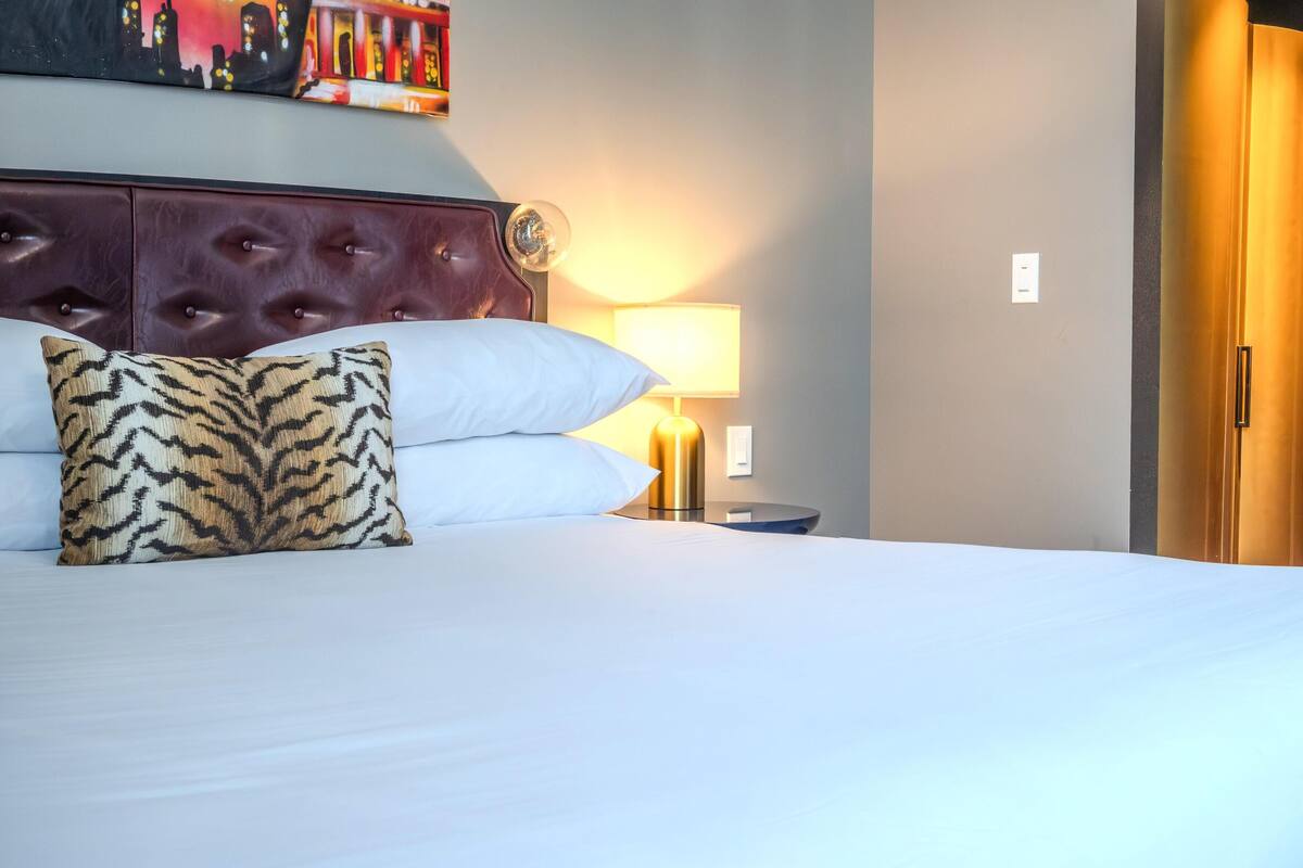 A hotel room with a tufted headboard, a bed with white linens, a tiger-patterned pillow, a bedside lamp, and a wardrobe.