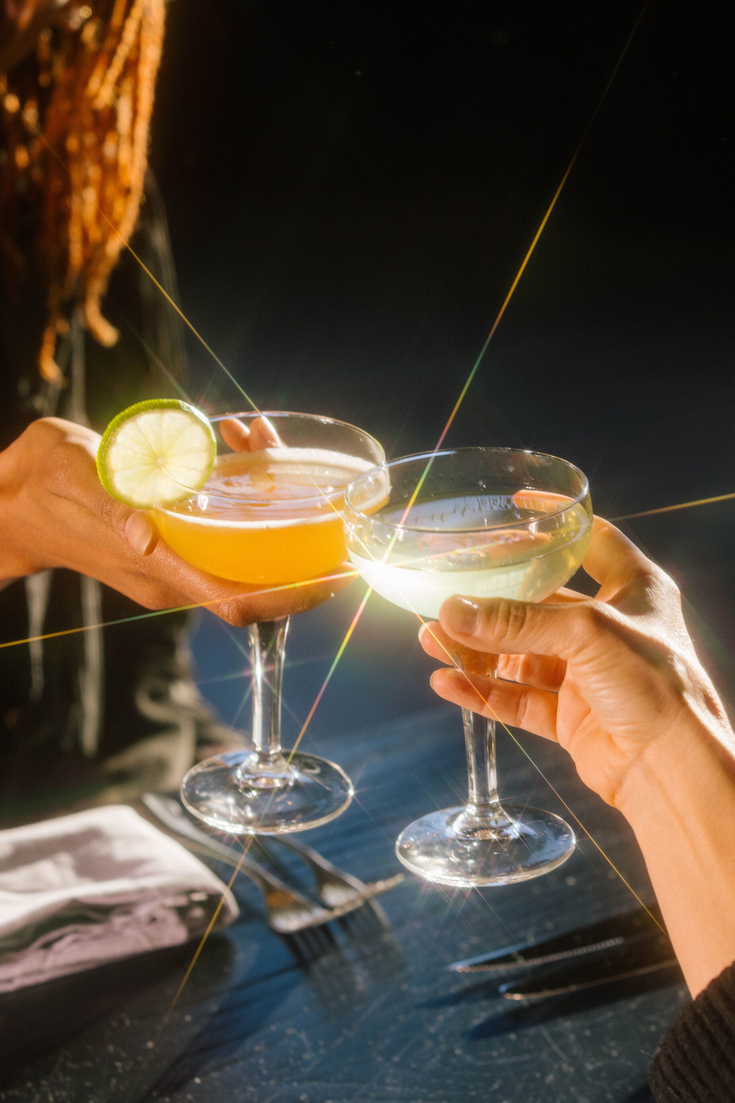 Zwei Hände halten Cocktailgläser mit Limettenscheibe, ein Löffel und eine Gabel liegen auf dem Tisch.