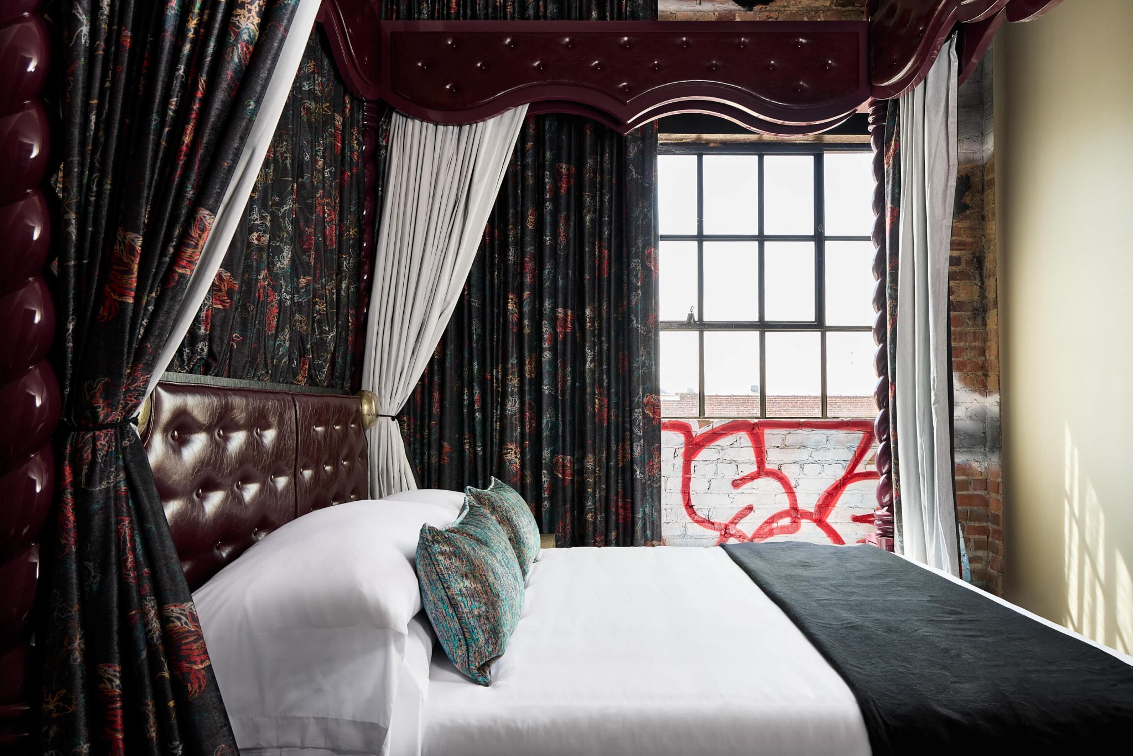 Ein Schlafzimmer mit einer gesteppten Kopfteil, einem Bett mit weißen Leinen und türkisfarbenen Kissen, dunklen Vorhängen mit Blumenmuster und Blick auf eine mit Graffiti versehene Backsteinmauer.