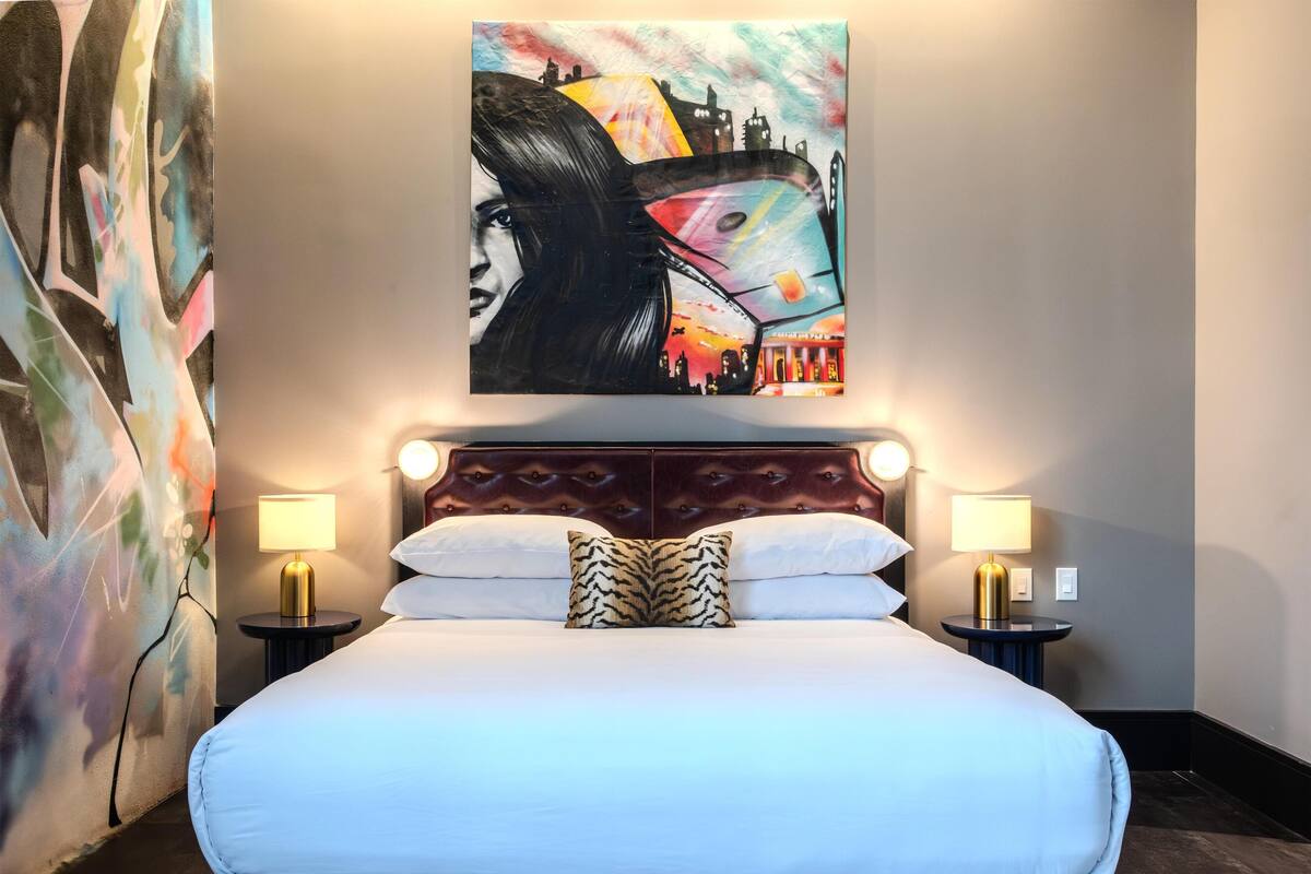 A hotel room with a large bed, two bedside lamps, a headboard with a light fixture, and a colorful abstract painting above the bed.